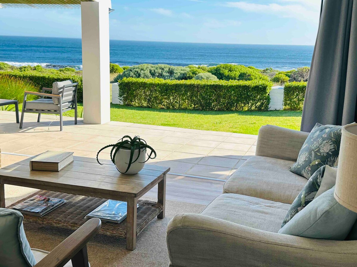 A comfortable living area features a neutral sofa and a rustic coffee table, set against a backdrop of ocean views. Large sliding glass doors open to a well-maintained garden, with greenery framing the expansive sea, inviting relaxation and outdoor enjoyment.