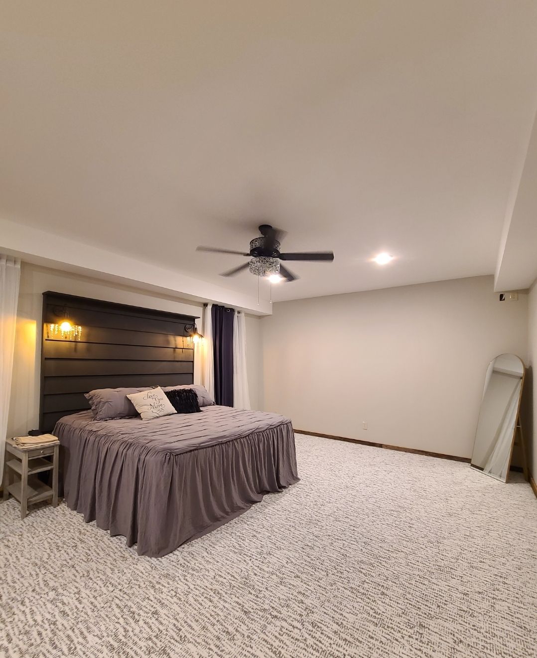A spacious bedroom features a plush, gray bedspread laid over a generous bed. Soft lighting is provided by wall sconces on either side. A decorative mirror stands against the wall, and the carpet has a subtle texture, enhancing the overall comfort of the space.