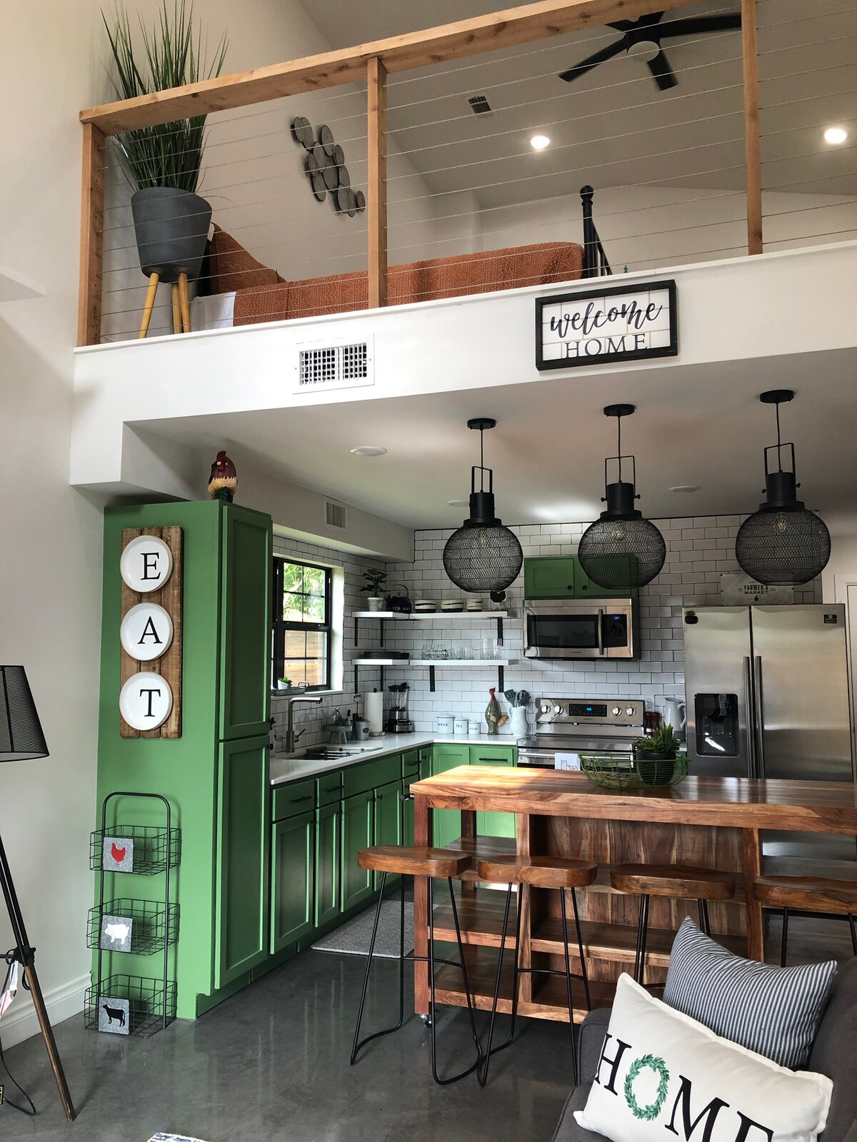 A modern kitchen is featured with green cabinetry and stainless steel appliances. A wooden dining table with chairs is positioned nearby. Decorative light fixtures hang above the countertop, while a small plant adds a touch of greenery. The upper level is visible with a welcoming sign.