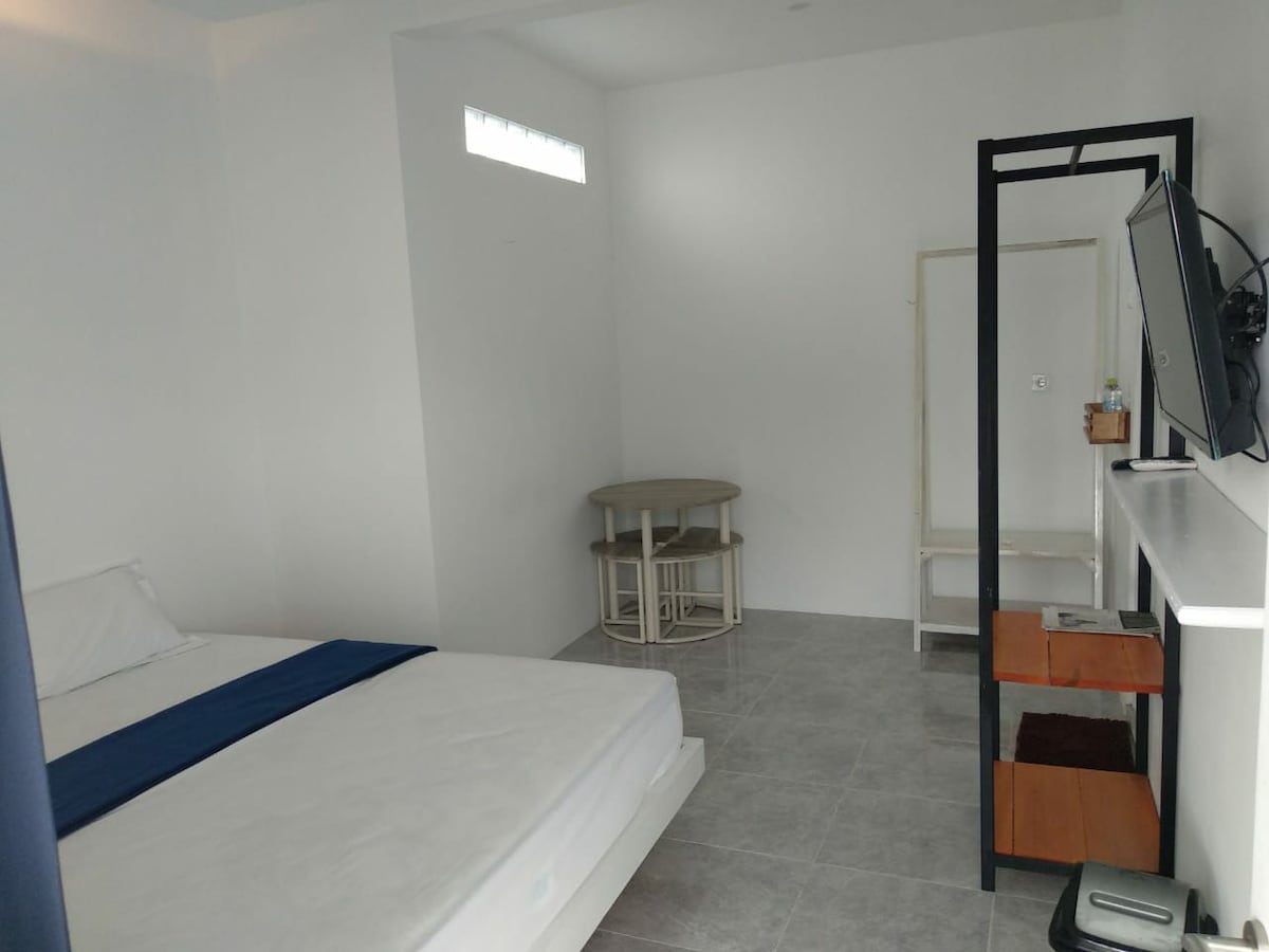 A bright and spacious room features a large bed with white bedding, complemented by a blue throw. A small table and two chairs are positioned nearby, while a minimalist wardrobe and a television are visible, enhancing the room's modern design.