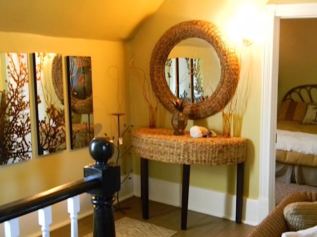 A charming entryway features a woven console table adorned with decorative items. A round mirror reflects the light, enhancing the welcoming atmosphere. Vibrant wall art in natural tones complements the yellow walls, while a glimpse of the adjoining bedroom is visible through an open doorway.