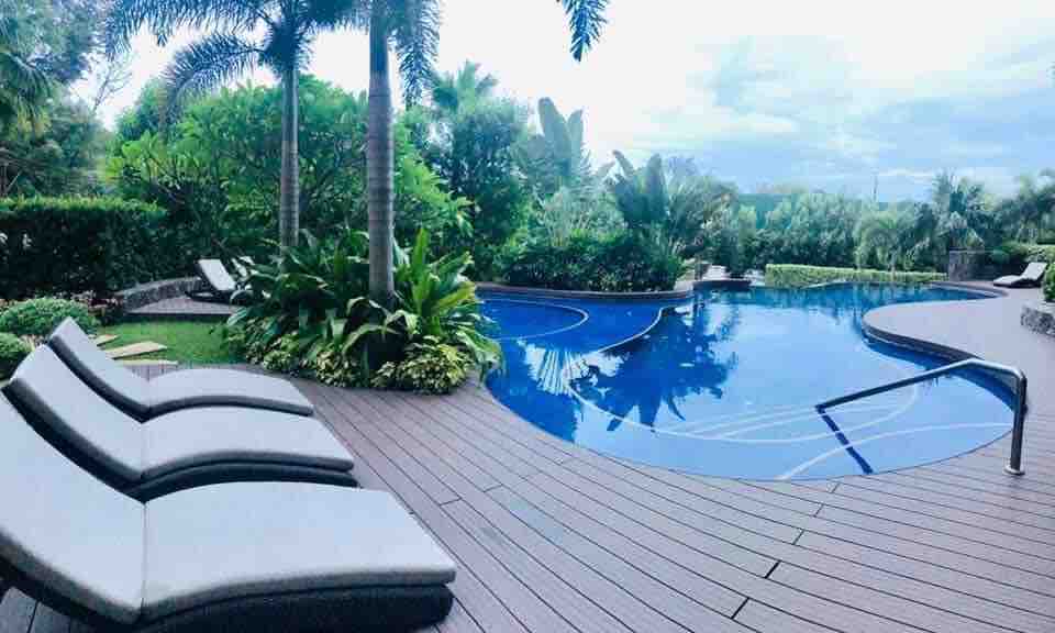 A serene pool area is surrounded by lush greenery, featuring a winding blue pool. Several lounge chairs are positioned nearby on an elegant wooden deck, creating a relaxing atmosphere. Tropical plants enhance the inviting outdoor space.