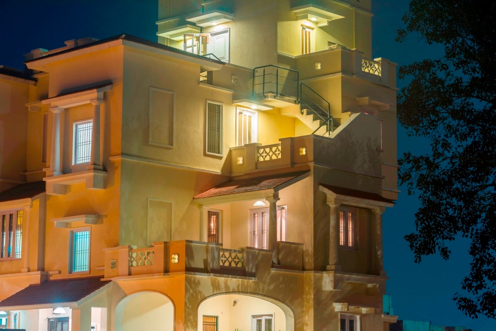 The exterior of the villa is highlighted by soft evening lighting, showcasing multiple levels with balconies and large windows. The architectural features include a terracotta roof and decorative railings, surrounded by lush trees that enhance the peaceful ambiance.