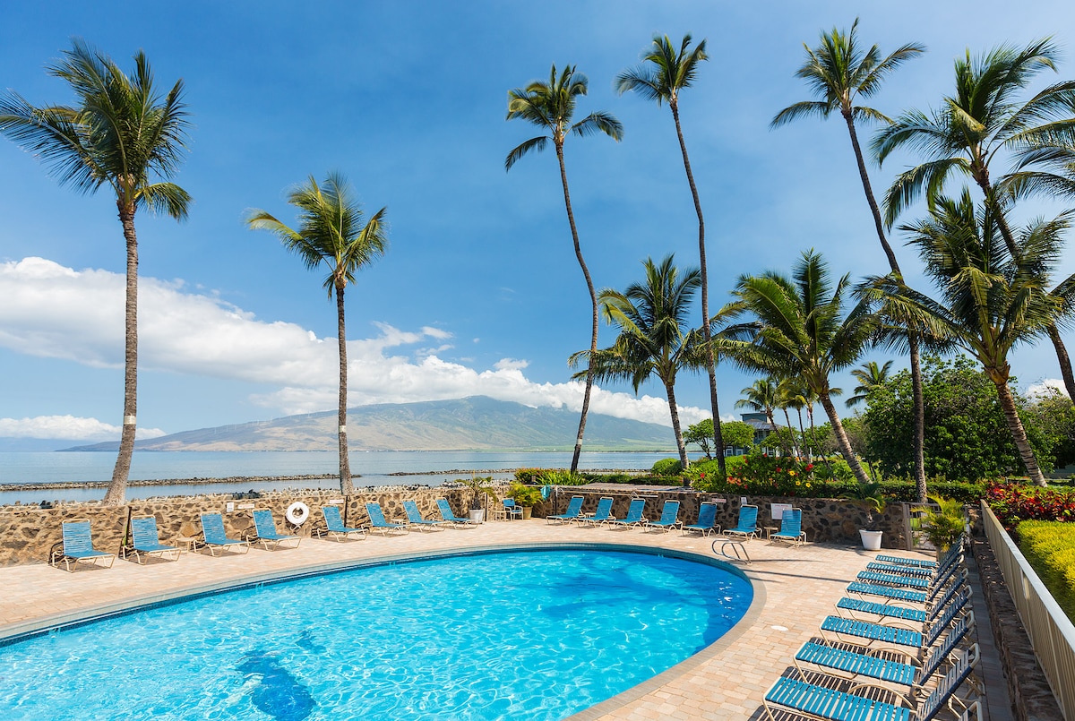 A large pool surrounded by palm trees is presented, with lounge chairs arranged along the edge. The clear blue water reflects the sky, while a scenic view of distant mountains and lush greenery is visible in the background.