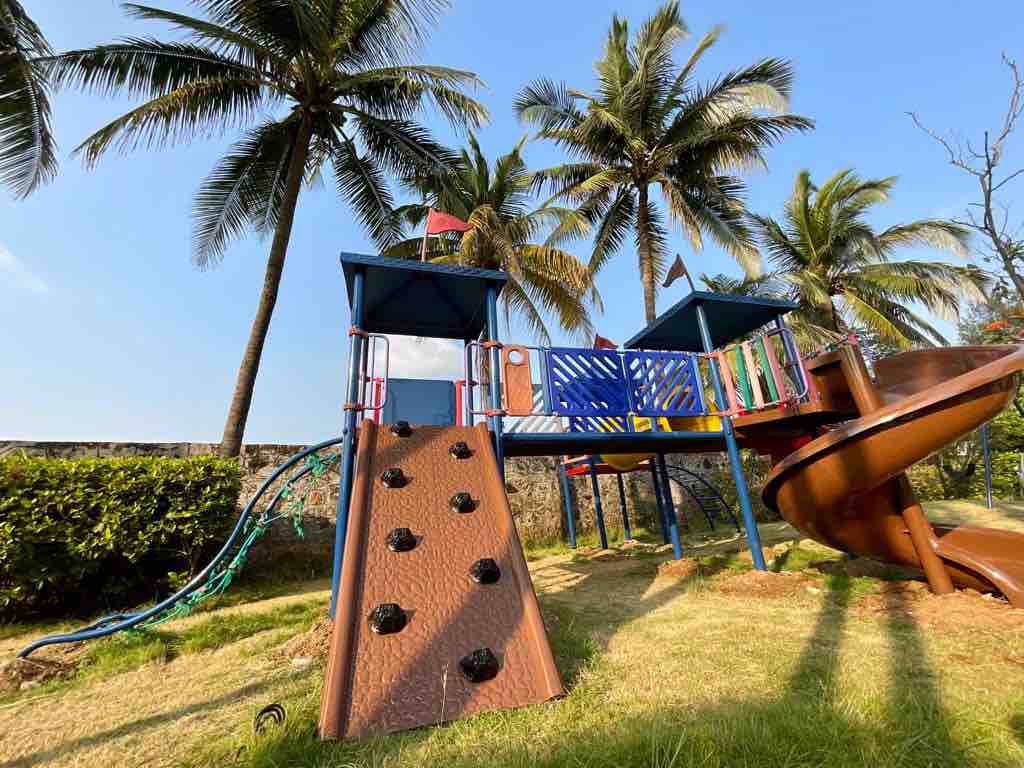 A colorful playground is positioned under palm trees, featuring a climbing wall, a slide, and various play structures. Green grass surrounds the area, offering a safe space for children to explore and play.