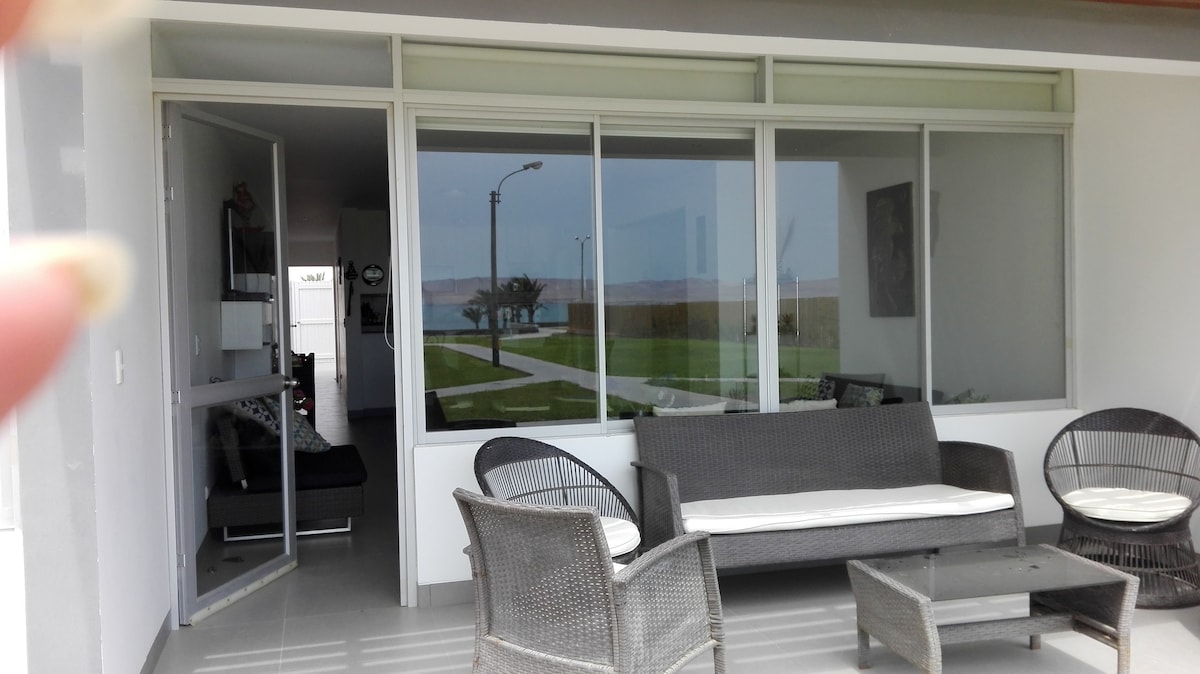 An inviting terrace area features a set of wicker furniture, including a coffee table and several chairs. Large glass windows provide views of the outdoor space and the distant sea, while the interior is partially visible, highlighting a cozy living area.