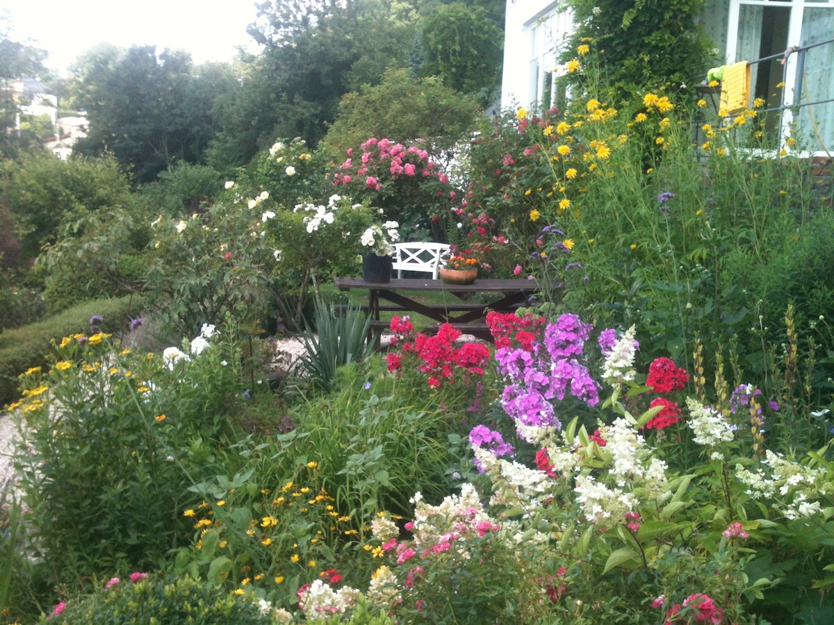 A vibrant garden is filled with an array of colorful flowers in various hues, including pink, white, yellow, and purple. A small white bench is nestled among the blooms, providing a serene spot to enjoy the natural surroundings.