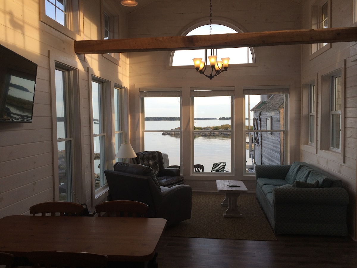 A bright living area showcases large windows offering unobstructed views of Moosabec Reach. Comfortable seating arrangements include a dark sofa and a light-colored couch, positioned facing the water. A wooden dining table and a light fixture enhance the space, inviting relaxation and tranquility.