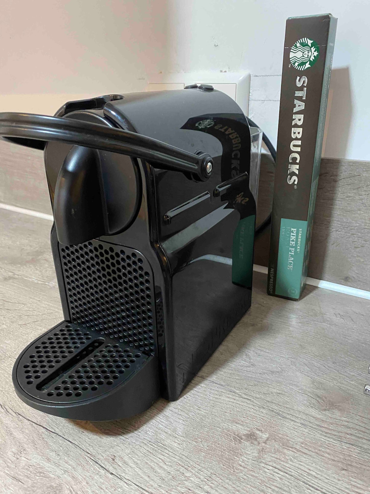 A sleek black coffee machine is positioned beside a box of Starbucks coffee pods. The machine features a simple design with a perforated drip tray and a side lever for easy operation.