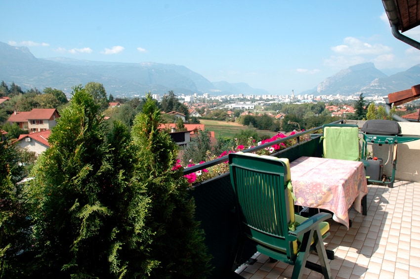 A spacious terrace is visible, featuring a table with floral tablecloth and two chairs, surrounded by greenery. In the background, breathtaking views of the surrounding mountains and the cityscape of Grenoble are presented, under a clear blue sky.