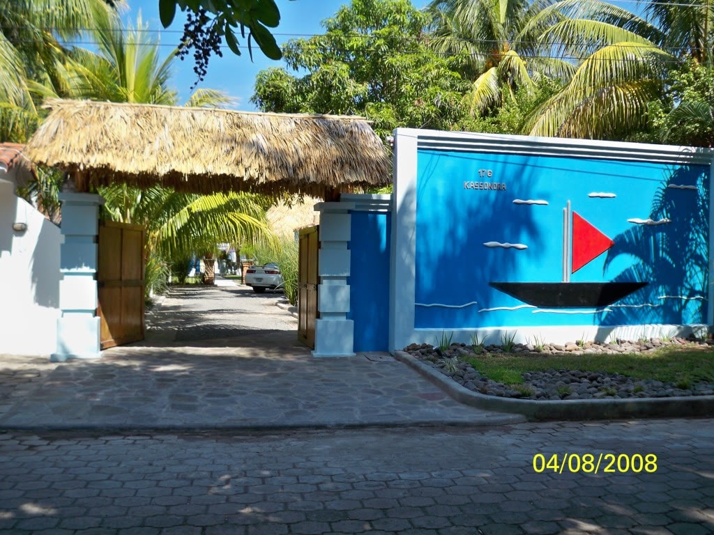 A vibrant entrance features a traditional thatched roof above wooden gates, complemented by a mural depicting a boat with a red sail against a bright blue background. Lush palm trees frame the entrance, creating a welcoming atmosphere to the property.