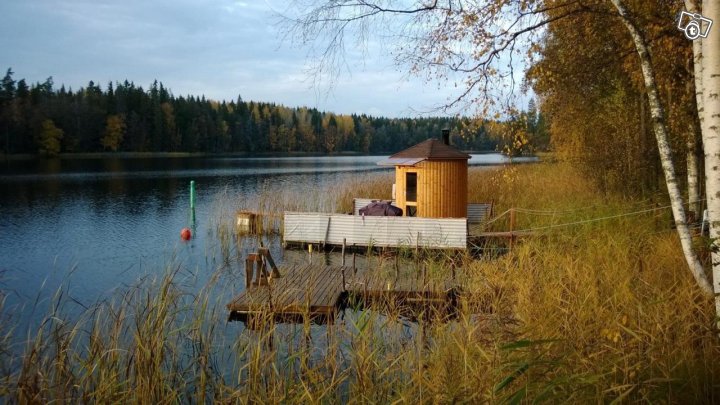 A wooden sauna is positioned on a floating dock by a calm lake, surrounded by tall grasses and autumn foliage. Soft ripples on the water reflect the natural scenery, while a buoy marks the shoreline nearby. The peaceful environment is complemented by the serene water setting.