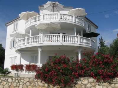 A three-story white villa is displayed, featuring multiple circular balconies adorned with white railings. Shade umbrellas are positioned above the balconies. Vibrant flowering plants in shades of pink and green surround the base of the building, creating a welcoming environment. A clear blue sky serves as the backdrop.