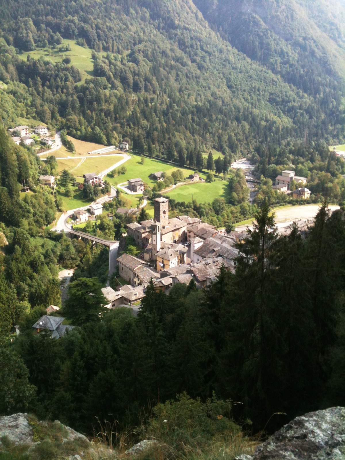 The medieval village of Gromo is nestled in a lush green valley, surrounded by mountains. Stone buildings and a prominent tower can be seen amidst trees, with winding roads leading into the village. A gentle stream flows through the area, enhancing the natural beauty.