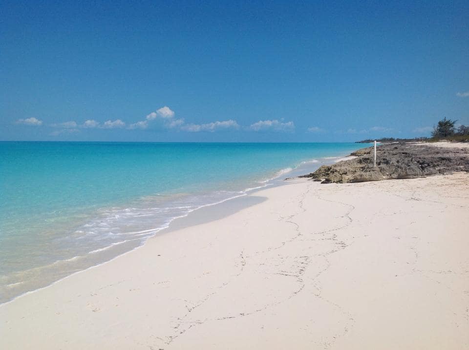A serene beach stretches along the coastline, featuring soft, white sand and gentle turquoise waves. The horizon showcases a clear blue sky with sparse clouds, while a rocky formation is visible along the shore, creating a natural boundary. Footprints trace the smooth sand.