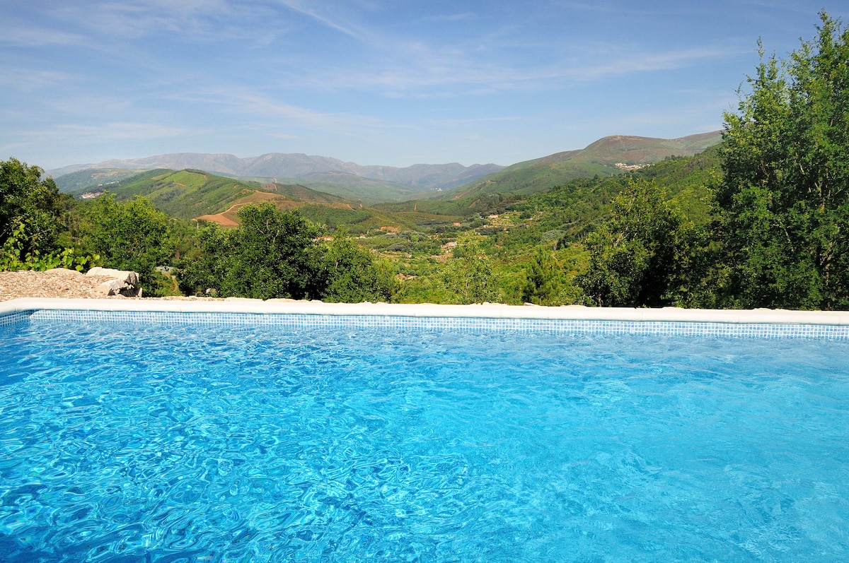 A private salt water swimming pool is edged by natural stone, offering clear turquoise water. The scenic view of rolling green hills and valleys stretches into the distance under a bright blue sky, adding a tranquil backdrop to the pool area.
