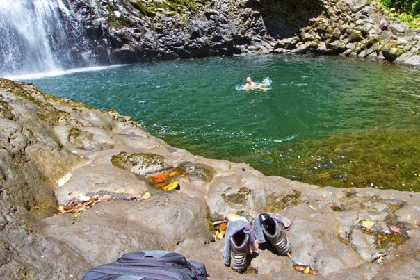 A serene natural pool is surrounded by rugged rocky terrain, with gentle water cascading over a nearby waterfall. A pair of shoes and a backpack are positioned near the pool's edge, while a person can be seen swimming in the refreshing water.