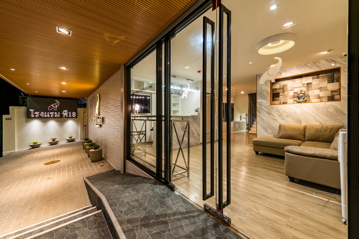 The entrance features spacious glass doors opening to a well-lit lobby. A sleek reception area and a comfortable seating arrangement create a welcoming environment. Decorative elements on the walls add a touch of design, complemented by modern lighting fixtures overhead.