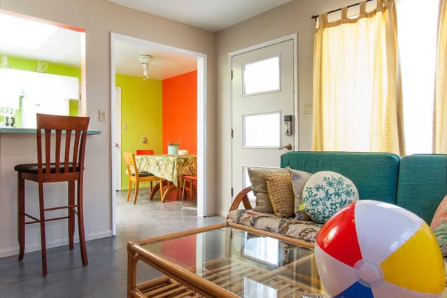 The living area features a cozy seating arrangement with a glass coffee table and colorful beach ball, enhancing the relaxed beach vibe. Light from nearby windows brightens the space, which flows into a dining area defined by bright green and orange walls.