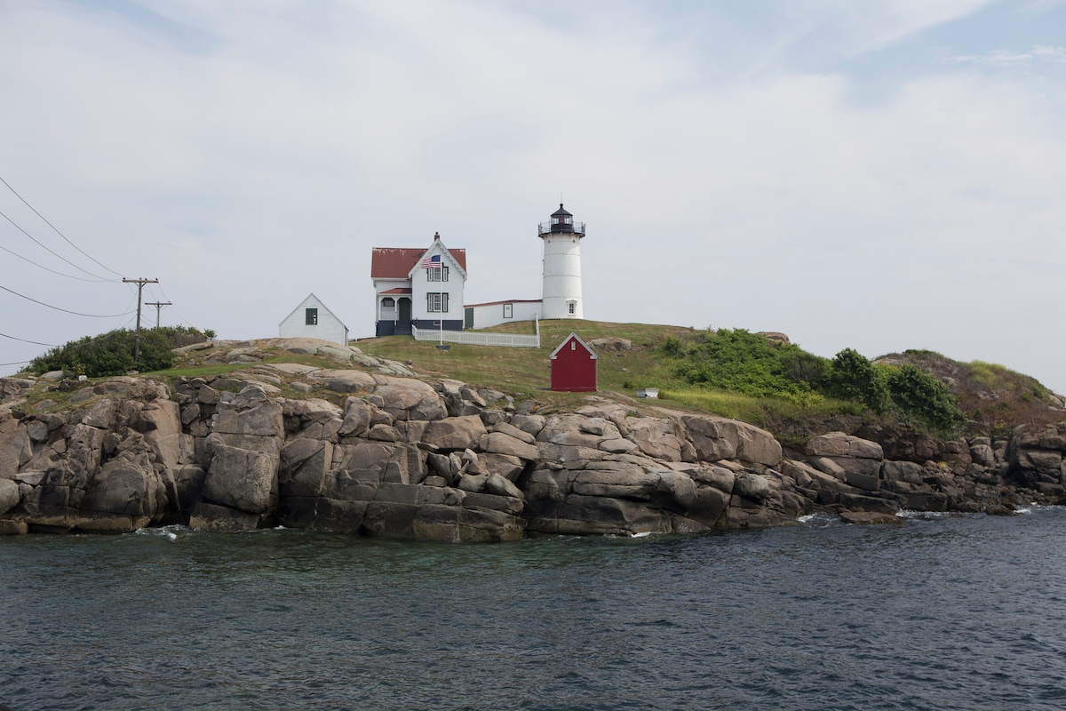 A historic lighthouse stands prominently on a rocky shore, surrounded by water. The white tower features a black top and is flanked by a white house with a red roof. A small red shed is visible in the background, adding a charming detail to the coastal scene.