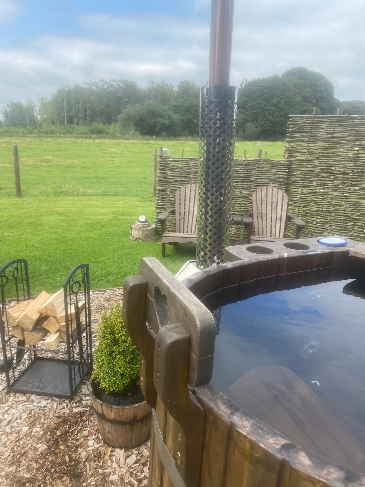 A wood-fired hot tub is placed in a scenic outdoor setting, surrounded by a grassy area. Seating is provided with two rustic chairs nearby, complemented by a wood storage area. Lush greenery borders the space, creating a peaceful atmosphere.