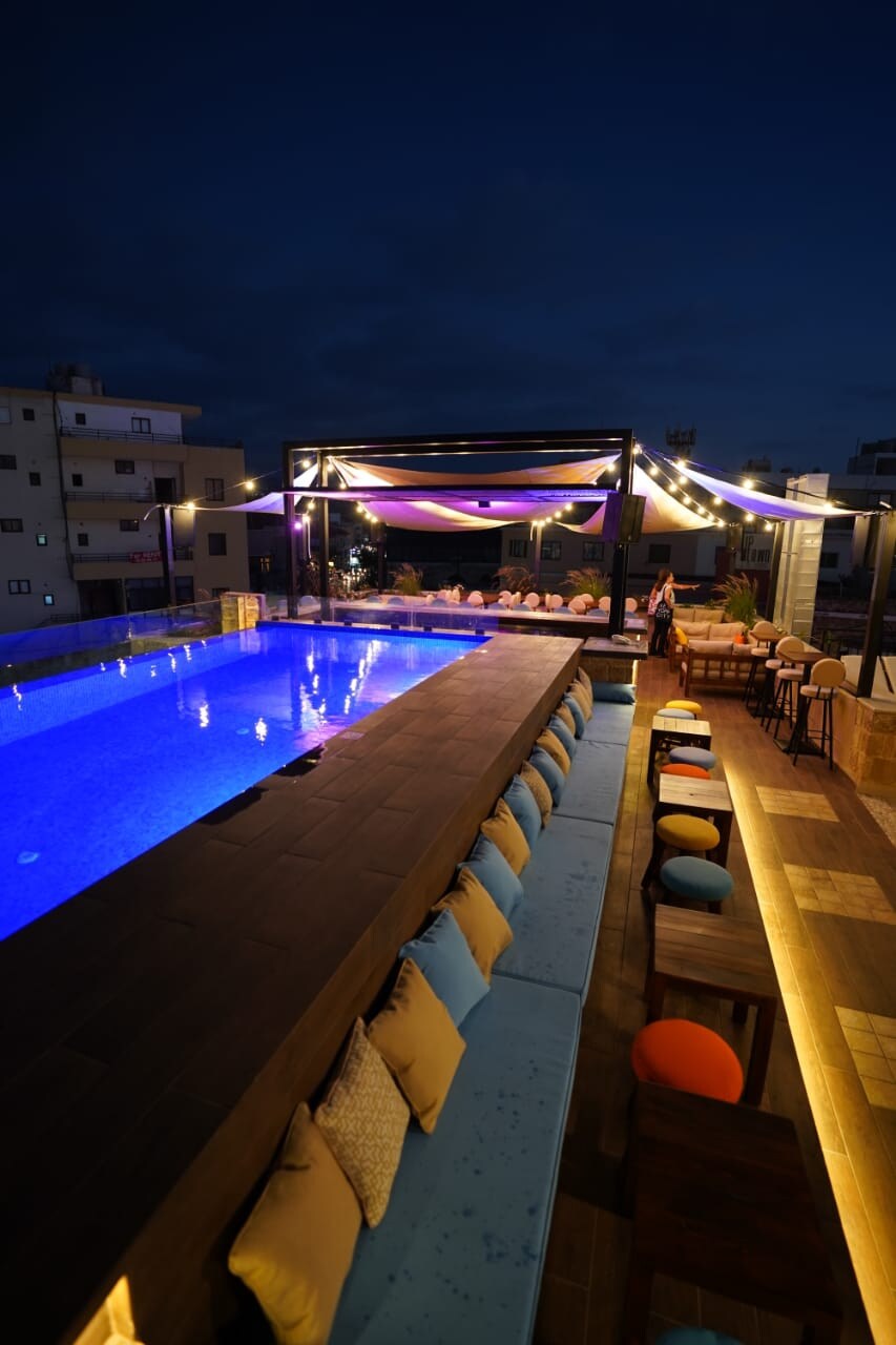 The rooftop lounge features a long bar beside a glowing blue swimming pool, with soft seating arranged along the edge. Warm lights create a welcoming ambiance against the night sky, while a variety of stools adds color to the space.