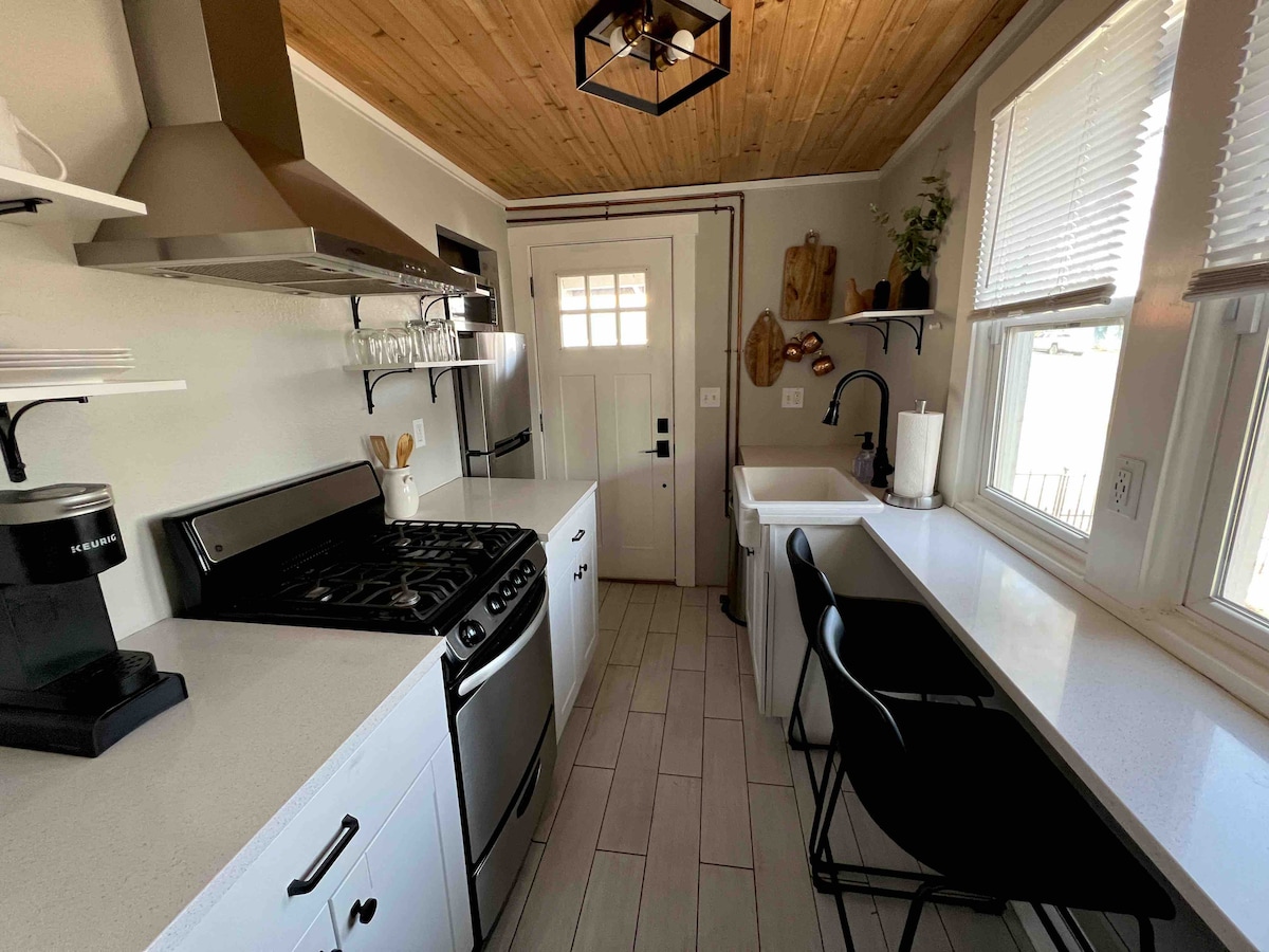 A modern kitchen is presented, featuring stainless steel appliances and a gas range. Light-colored cabinets and countertops create a bright environment. A wooden ceiling adds warmth. A sleek black faucet is positioned above a white sink, with a small table and two black chairs arranged along the side.