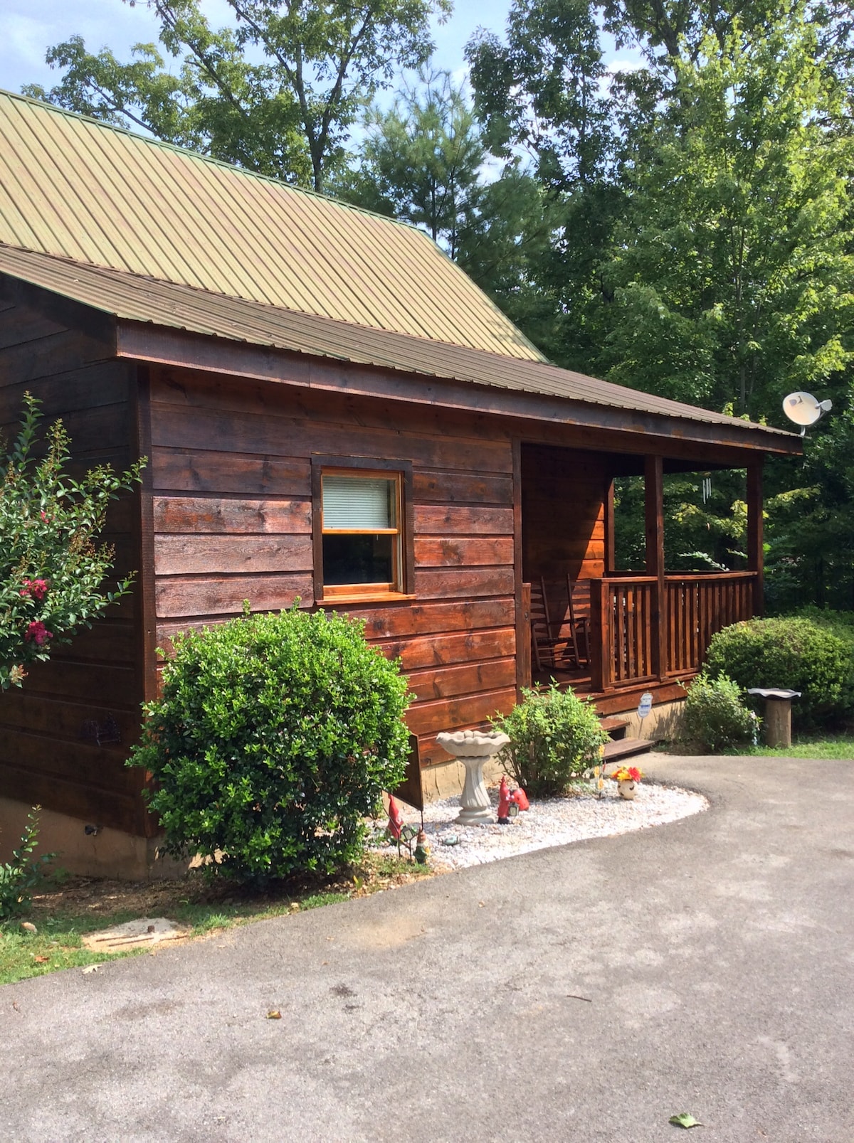 A charming wooden cabin is nestled among greenery, featuring a sloped metal roof and a covered porch with wooden railings. The entrance is framed by small shrubs and decorative garden elements, conveying a welcoming outdoor space.