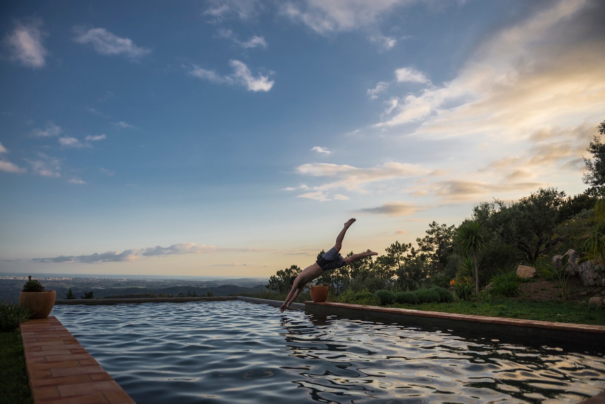 A person is captured diving into a private infinity pool surrounded by lush greenery. The tranquil water reflects the sky, where clouds blend softly with warm sunset hues. The serene landscape provides expansive views of the Algarve in the background.