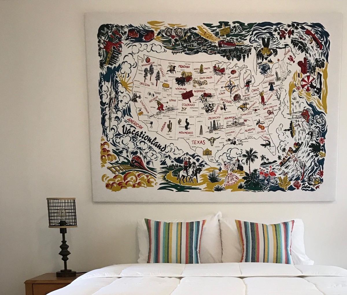 A spacious bed is positioned against a colorful map artwork of the United States, featuring illustrations of various states and landmarks. Two striped accent pillows provide a vibrant contrast, while a small lamp stands on a wooden nightstand to the side.
