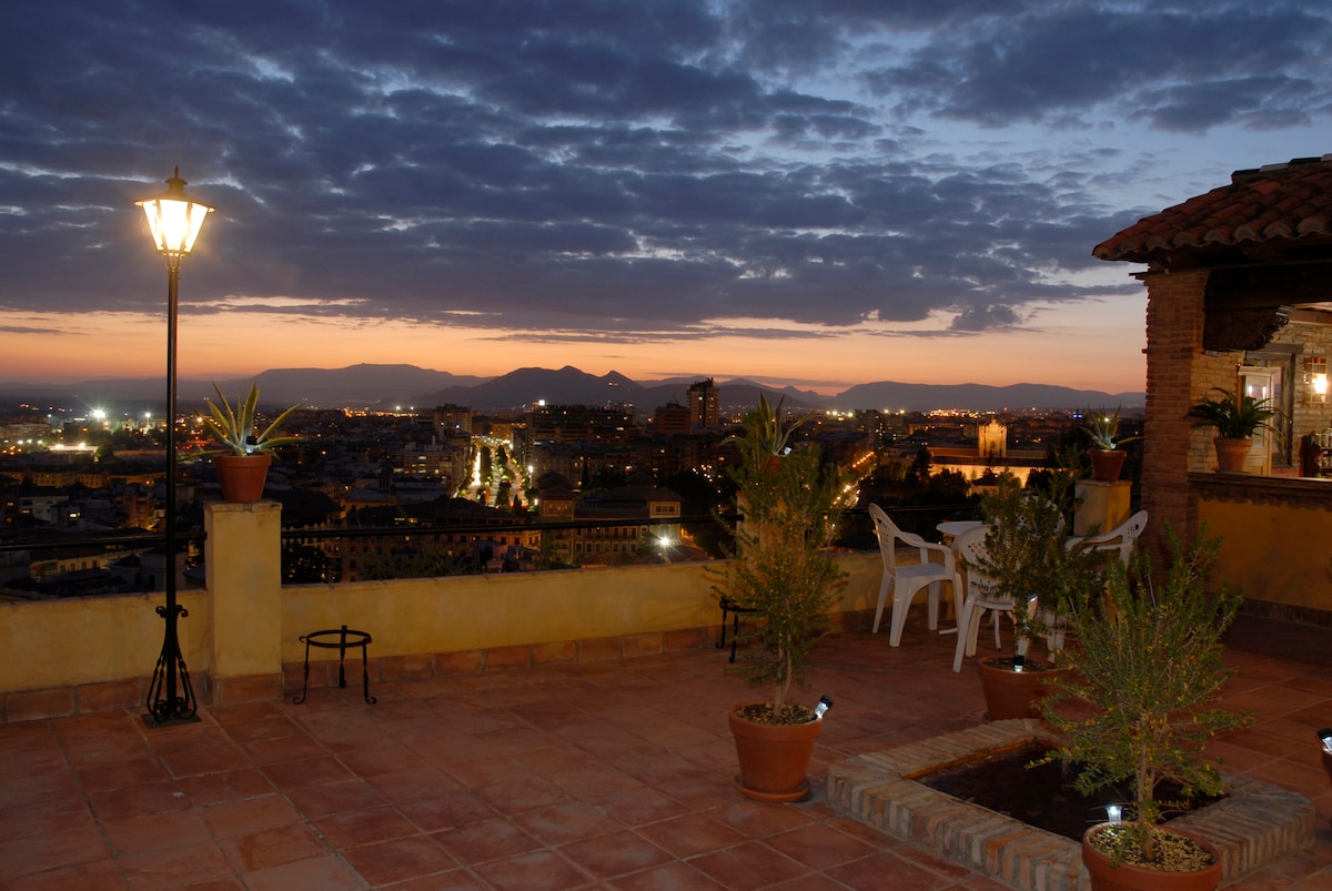 A spacious terrace showcases a scenic view of the city at sunset. Potted plants are arranged thoughtfully, and a table with chairs is positioned to maximize the panoramic sight. A lamp provides soft illumination, while the distant mountains frame the horizon under a twilight sky.