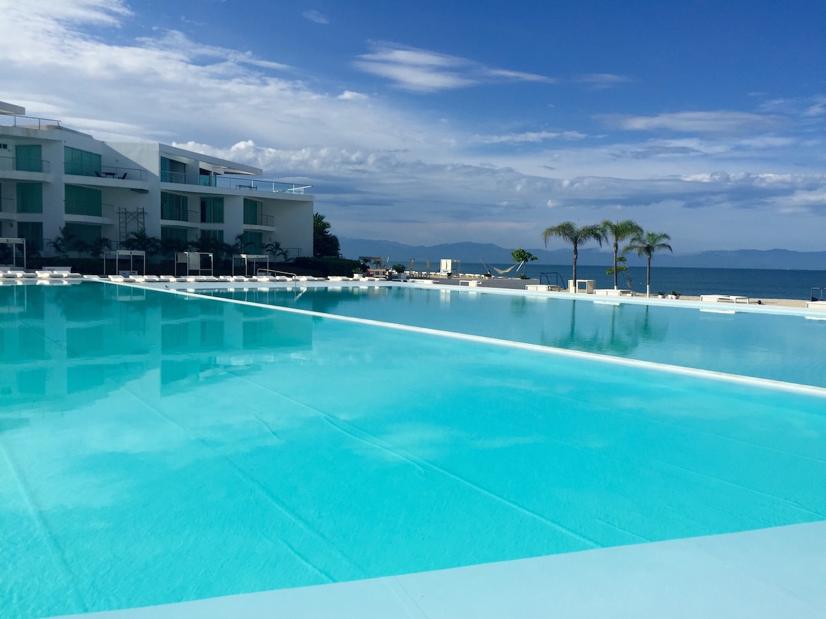 A large swimming pool is featured, displaying clear blue water under a bright sky. The pool area is surrounded by lounge chairs and palm trees, with a view of the ocean and distant mountains in the background.