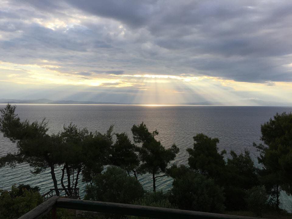 A scenic view of the sea is presented under a cloudy sky, with rays of sunlight breaking through clouds and reflecting on the calm water. Sparse trees frame the foreground, highlighting the tranquility of the coastal landscape.