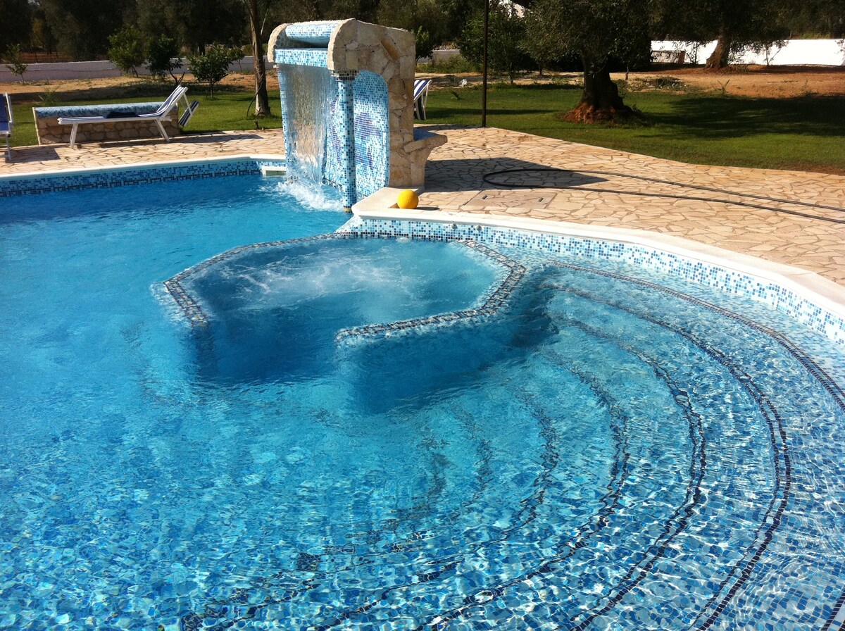 A tranquil pool area is featured, highlighting a shimmering blue pool with integrated hydro-massage and a waterfall. Comfortable lounge chairs are arranged nearby, surrounded by lush green grass and olive trees, creating a serene outdoor atmosphere.