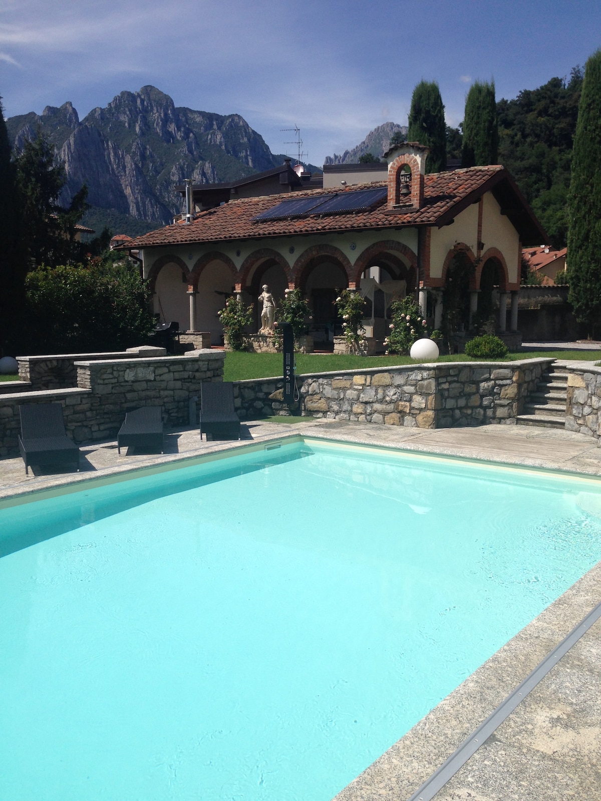 A clear swimming pool reflects the sky, surrounded by stone tiles. Loungers are arranged neatly beside the pool. In the background, a charming building with arched entryways and a tiled roof is set against a mountain backdrop, adding to the scenic landscape.