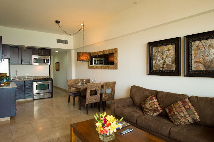 A modern living area is showcased, featuring a comfortable sofa adorned with patterned cushions. A wooden coffee table sits in front, accompanied by a dining table with woven chairs. The kitchen area is equipped with stainless steel appliances, and decorative artwork is displayed on the walls.