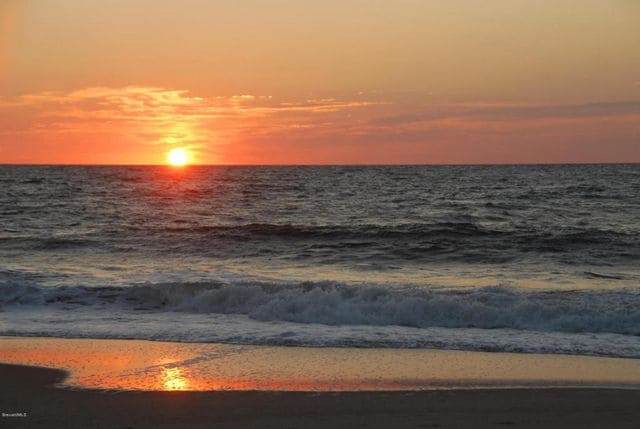 The ocean horizon presents a serene sunset, with the sun gradually descending, casting warm orange and pink hues across the sky. Gentle waves reflect the colors on the sandy beach, creating a tranquil scene that invites relaxation.