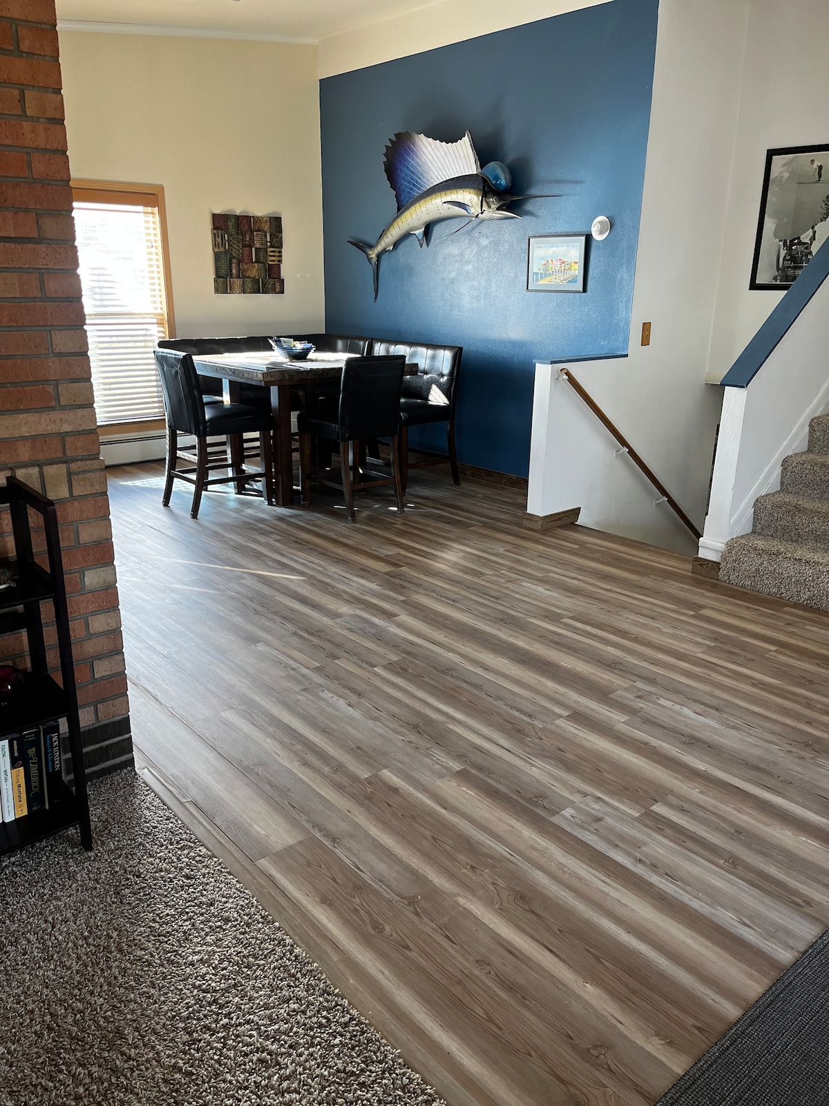 A spacious entryway is highlighted by wood-look flooring that guides towards a dining area featuring a round table surrounded by dark chairs. A blue accent wall displays a decorative fish mount, while a staircase leads to the upper level.