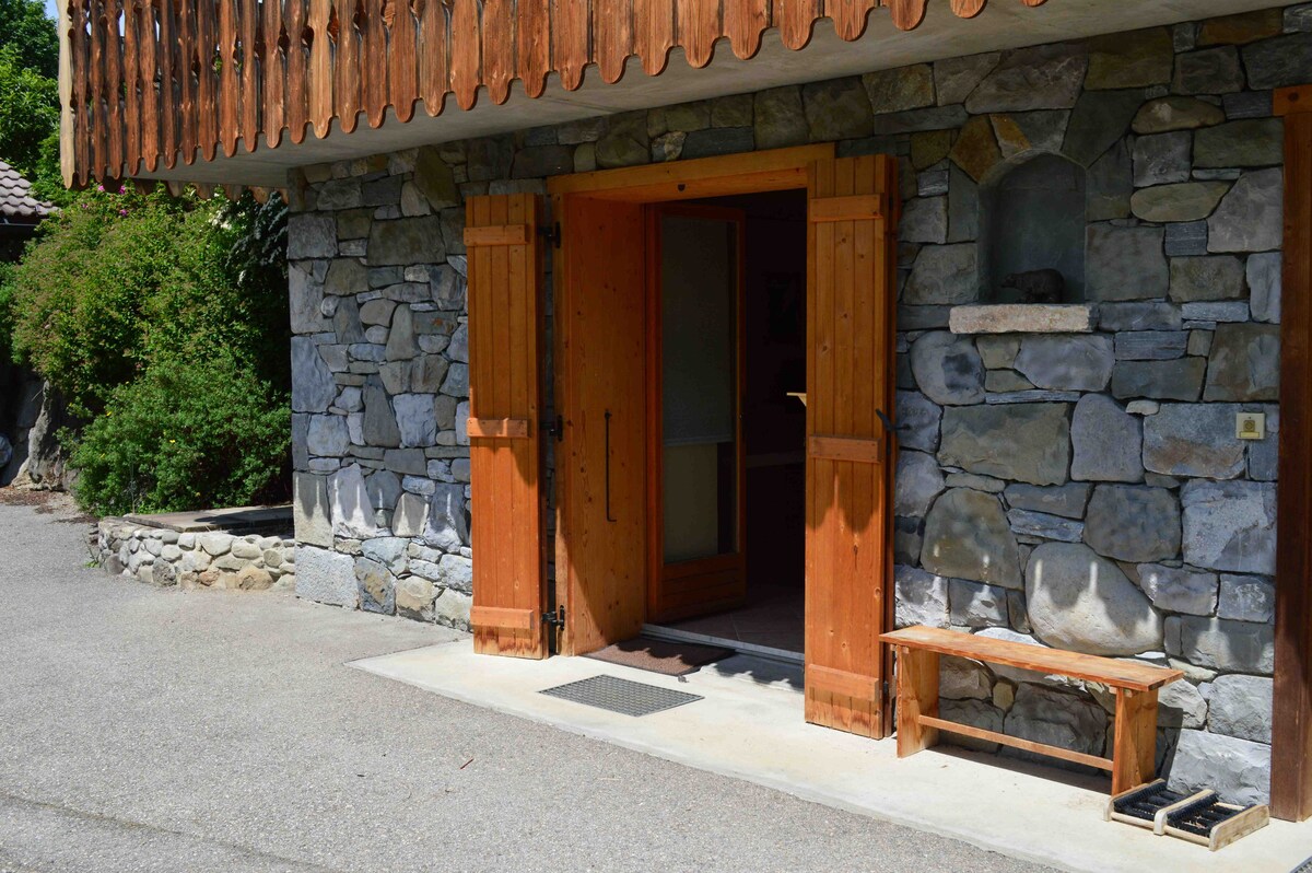 The entrance of the stone chalet features wooden double doors with shutters. A simple wooden bench is placed beside the door. The setting includes a landscaped area with greenery, complementing the rustic charm of the structure.