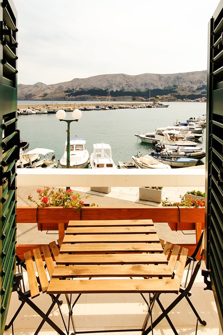 A view from an open balcony showcases the marina, with several boats bobbing gently in the water. A small wooden table and chairs are positioned on the balcony, framed by green shutters and colorful flowers in planters.