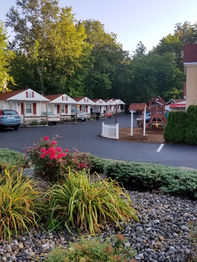 A line of charming cabins is visible, each featuring bright red roofs. A well-maintained playground is seen in the foreground, surrounded by vibrant flower beds and landscaped greenery, creating a welcoming atmosphere. Ample parking space is also present.