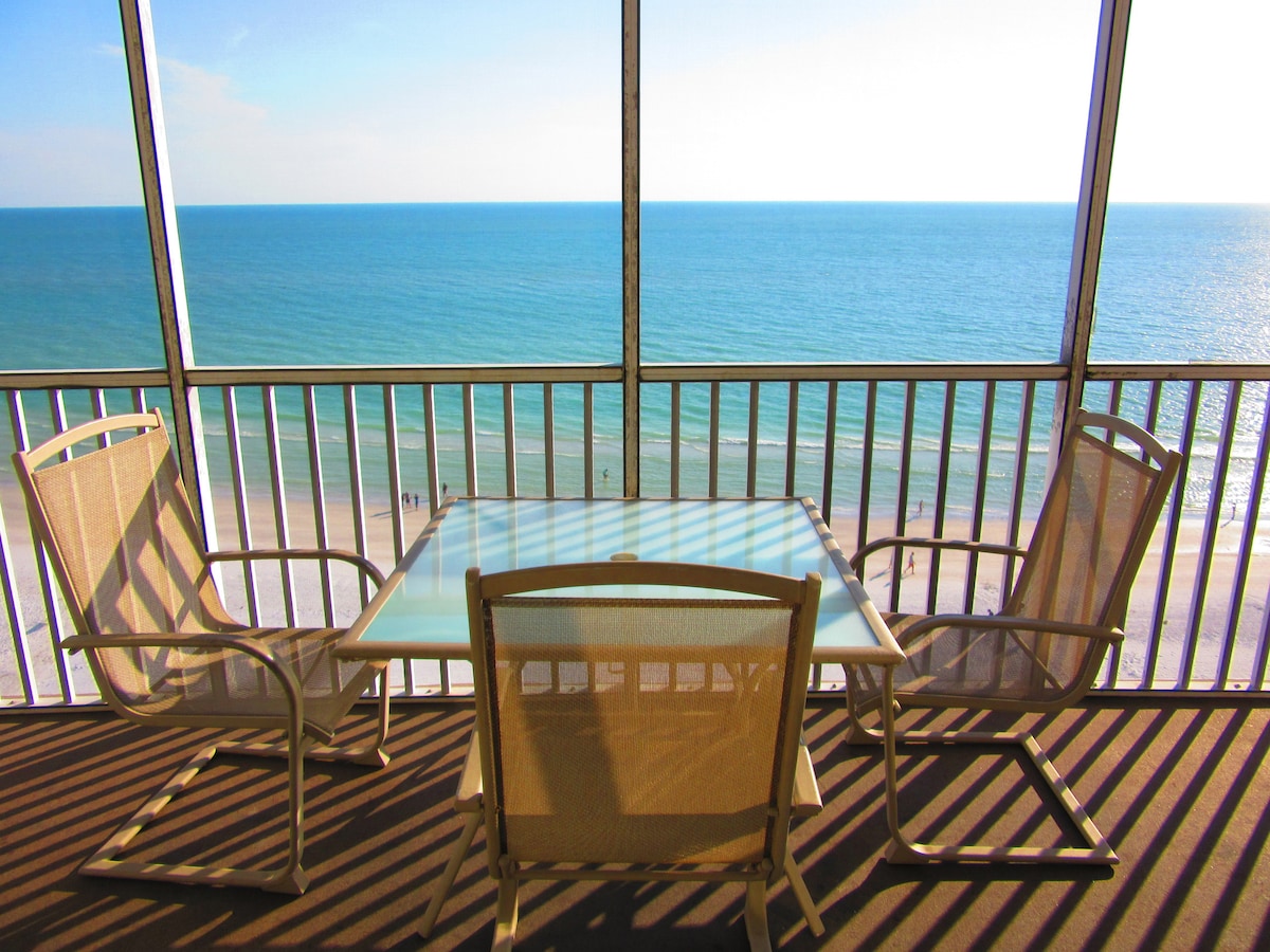 A screened lanai features a glass-top table surrounded by four beige chairs, offering unobstructed views of the Gulf. The sun reflects off the tranquil water, creating a serene atmosphere. Light shadows cast from the railing pattern enhance the inviting space.