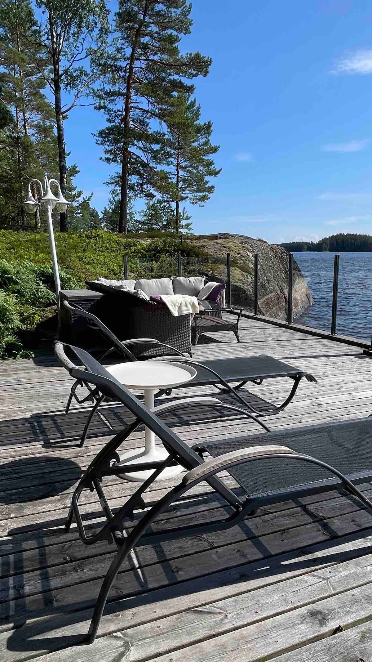 The outdoor area features a wooden deck alongside a tranquil lake, equipped with lounge chairs and a small table. A comfortable seating arrangement is visible with a blanket draped over a chair. Soft greenery and pine trees frame the scenic view of the water.