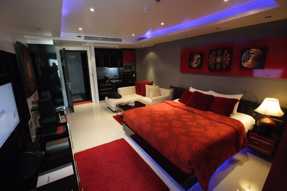 The luxurious suite features a spacious bed with red bedding, complemented by dark wood furnishings. A white sofa set is present, along with a sleek kitchen area. Decorative wall art and ambient lighting enhance the modern design, while a large flat-screen TV is visible.