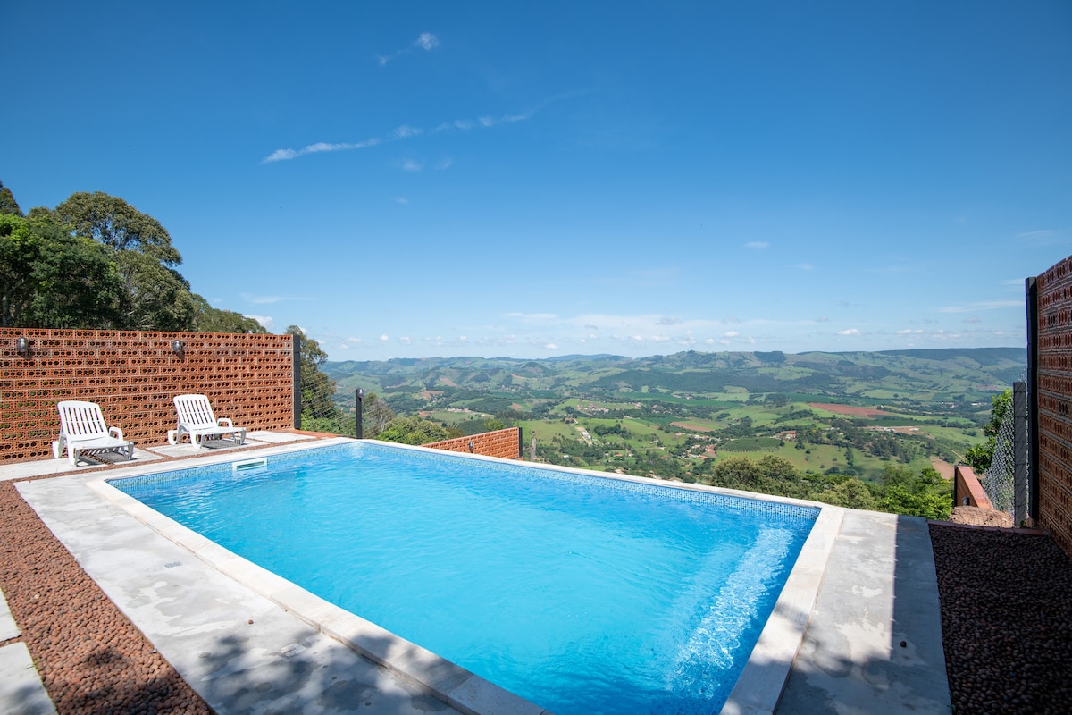 A rectangular swimming pool features clear blue water, surrounded by a stone deck and two white lounge chairs. A scenic view of rolling green hills and valleys extends in the background under a bright blue sky.