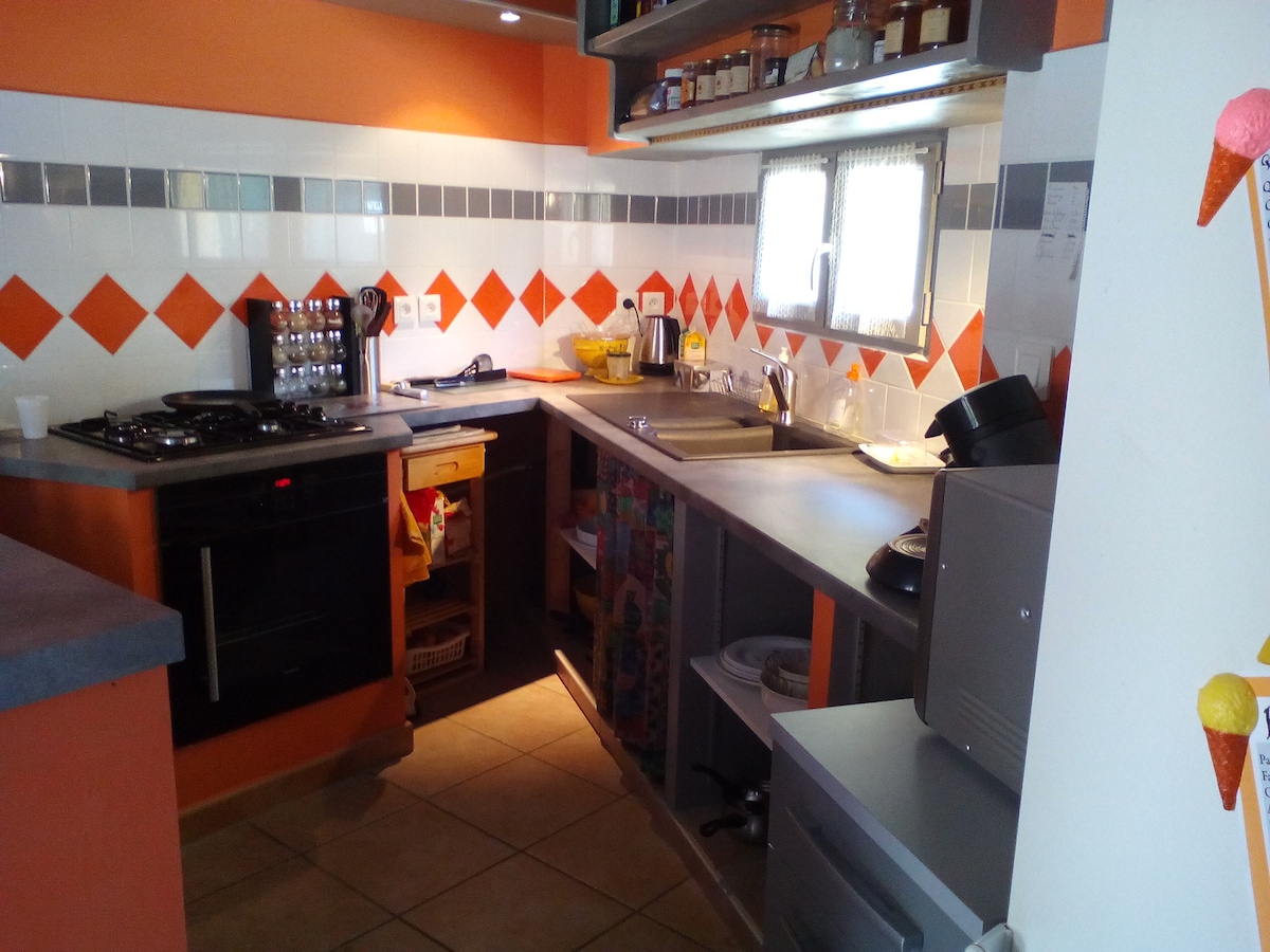 An equipped kitchen features a stove, oven, and various storage cabinets. Bright orange wall accents contrast with neutral tiles and countertops. Shelves display jars and utensils, while a window provides natural light. The layout promotes functionality with ample counter space for meal preparation.