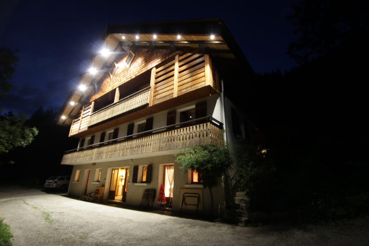 The exterior of a chalet is illuminated at night, showcasing wooden balconies and a warm glow from the ground floor windows. A gravel driveway leads up to the entrance, with parked vehicles nearby and surrounding greenery enhancing the peaceful environment.