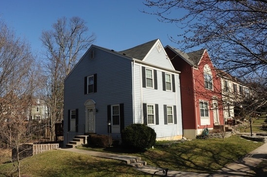 A two-story townhouse is viewed from the side, featuring a light blue exterior with black shutters. Steps lead to a white front door, while a small landscaped area with grass and shrubs is visible in front. A neighboring red townhouse is also shown in the frame.