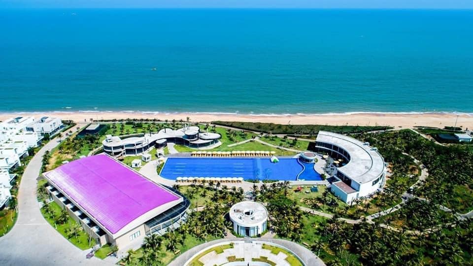 An aerial view captures the expansive Oceanami Resort, showcasing a large swimming pool surrounded by palm trees. The coastline can be seen in the background, along with the blue sea under a clear sky. Various buildings are arranged thoughtfully across the property.