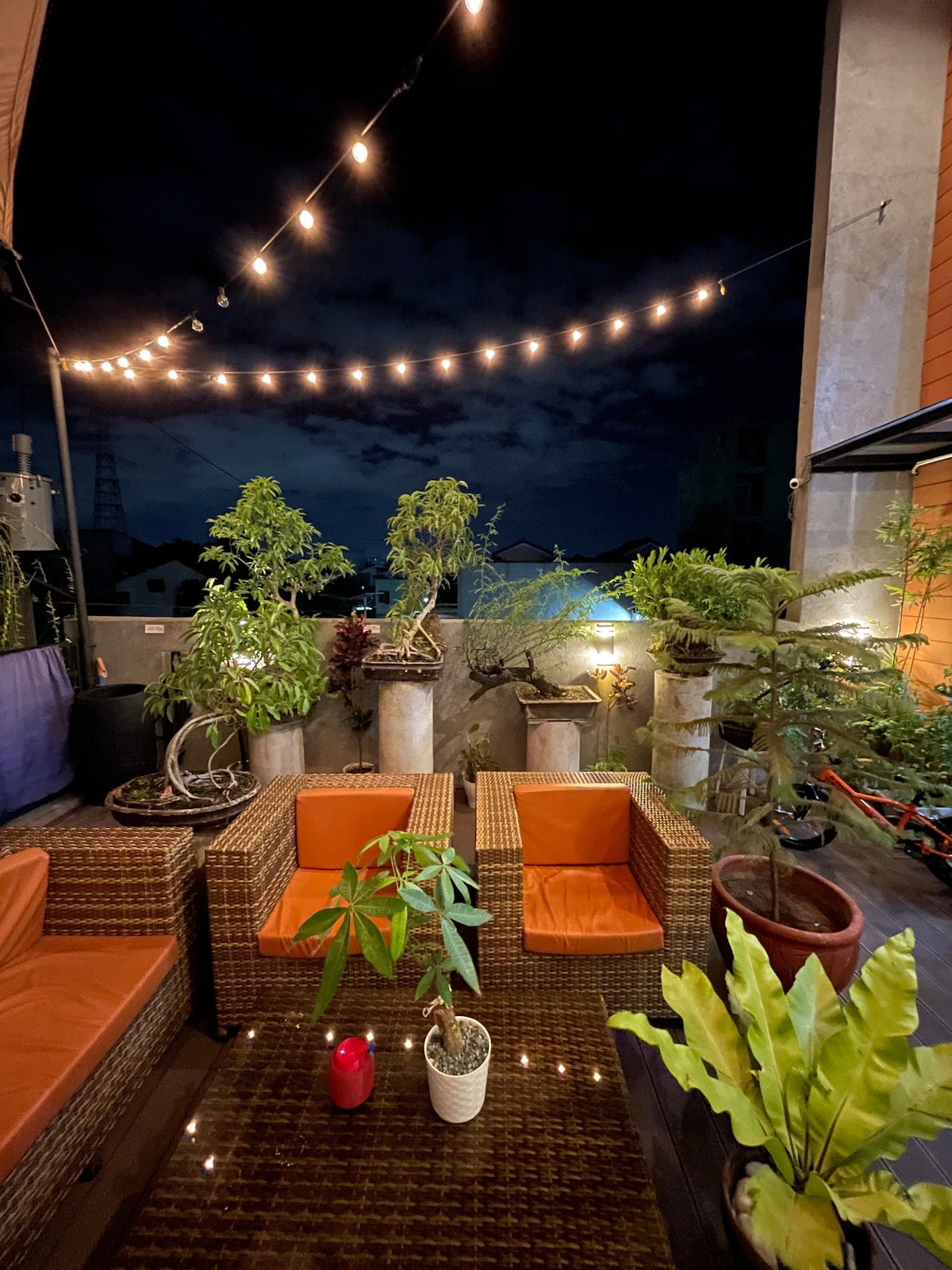 A cozy outdoor seating area is surrounded by potted plants and illuminated by string lights, creating a tranquil evening ambiance. Two rattan chairs with orange cushions face a glass-top coffee table, while various greenery adds a touch of nature. The backdrop features a darkening sky and distant city views.